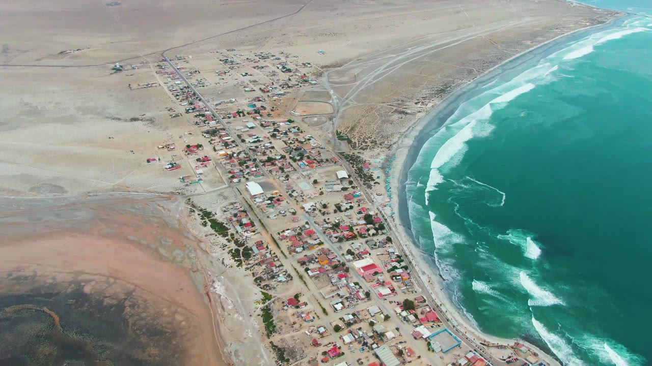 Wide aerial frame over Punta Abreojos showing a small road-lined village between sandy desert terrain and a curved turquoise shoreline, with no visible people.
