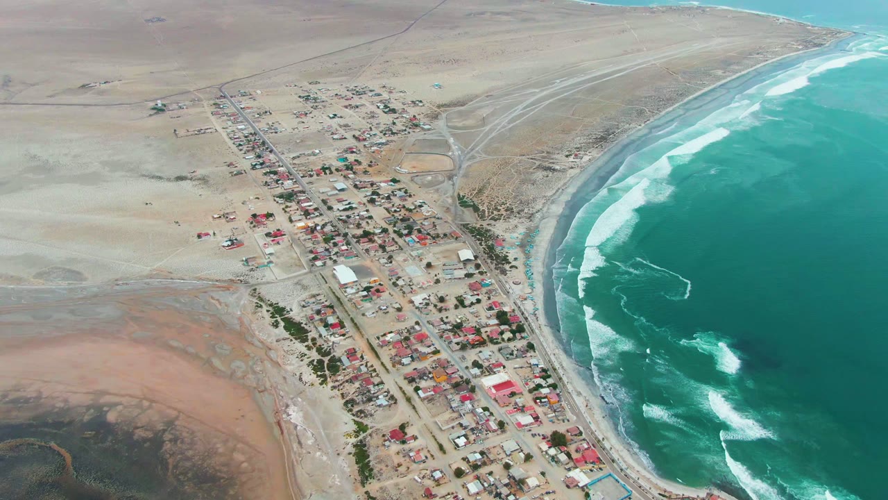Coastal settlement, sandy terrain, curved beach, and breaking ocean waves from above.