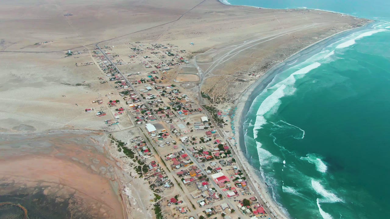 Punta Abreojos appears from above with the settlement centered lower in frame, open sandy land behind it, and bright turquoise water filling the right edge, with no visible people.