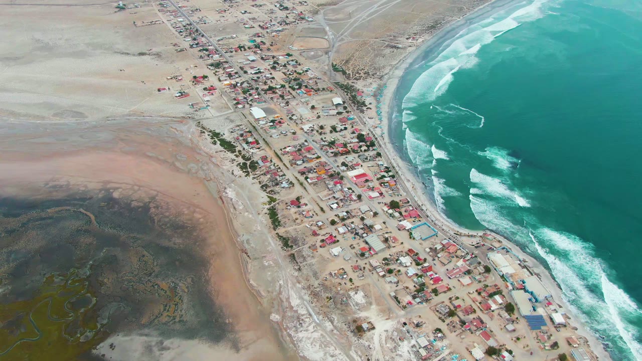 High aerial view of Punta Abreojos, Baja California Sur, with a compact coastal village between pale dry terrain and turquoise ocean, no visible people.