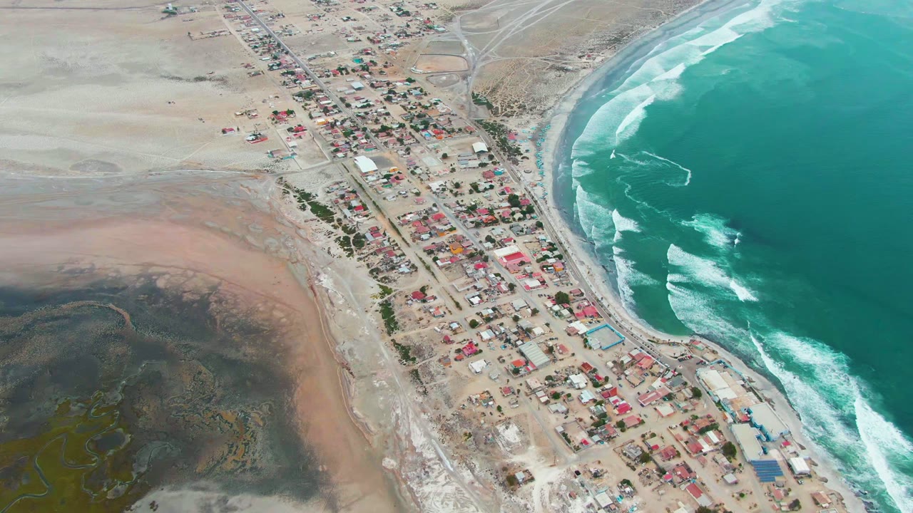 Oblique drone view of shore buildings, beach, and white surf.