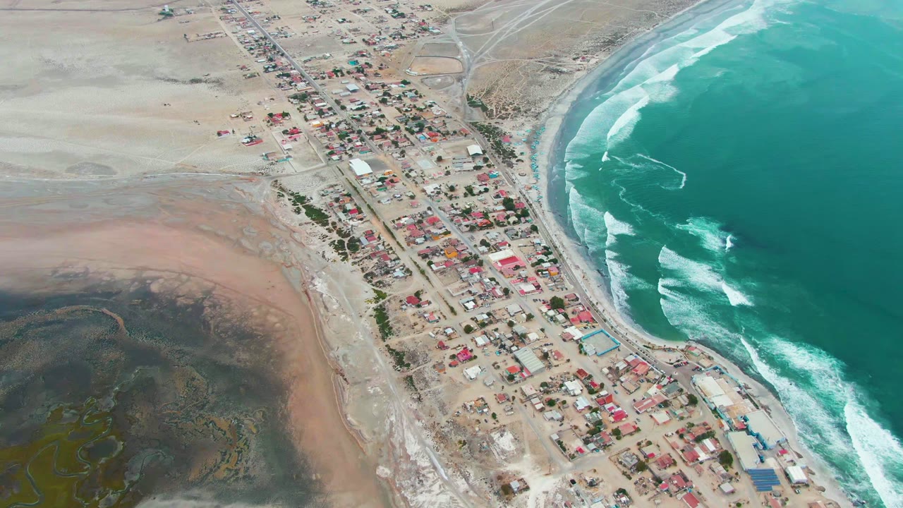 Aerial frame of Punta Abreojos with the village centered between broad sandy flats on the left and curved ocean waves on the right, no visible people.
