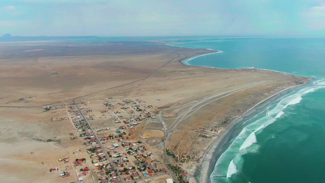 Opening aerial frame over Punta Abreojos, Baja California Sur, with a small settlement and straight road near a curving beach, white surf on the right, desert stretching inland, and no visible people.