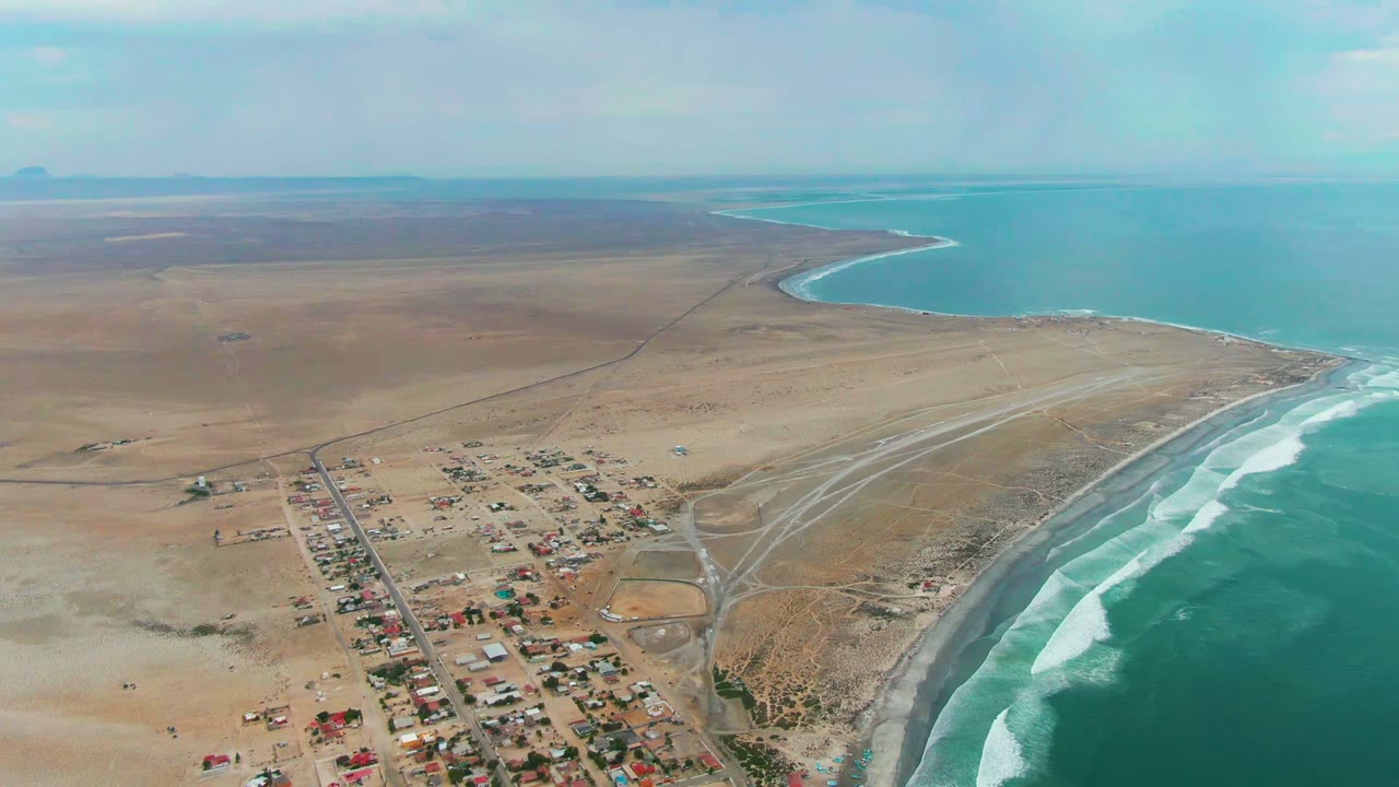 Wide drone view of desert settlement, beach roads, and breaking waves.