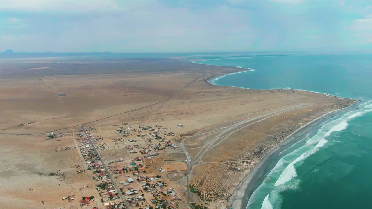 Mid-clip aerial frame of Punta Abreojos with clustered rooftops near the lower center, a branching sandy road pattern by the beach, bright ocean water on the right, and no visible people.