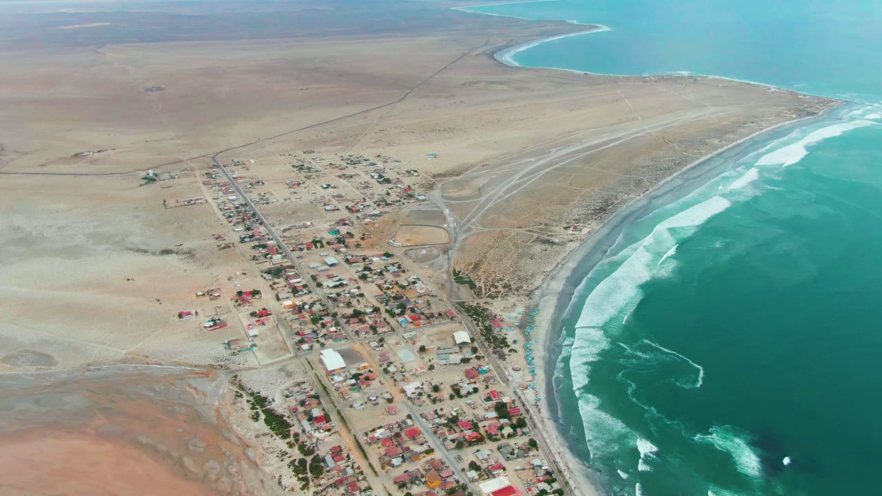Aerial view of Punta Abreojos, Baja California Sur, Mexico, with small buildings and roads near a curved surf line, beige desert beyond, and no visible people.