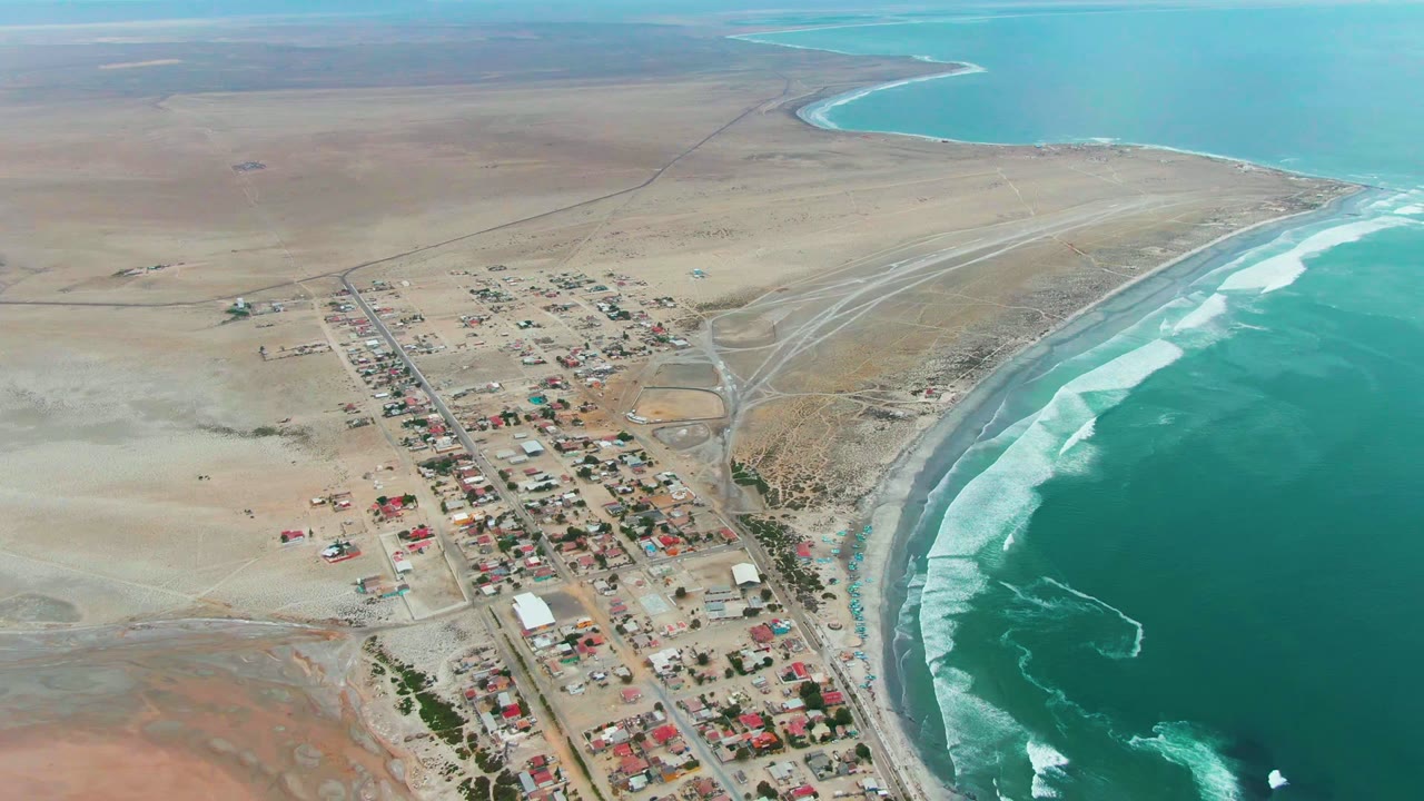 High aerial view of desert town roads beside white surf and blue-green ocean.