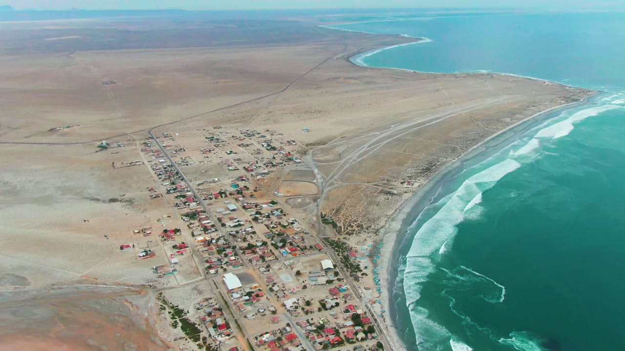 Aerial view of Punta Abreojos with the settlement centered lower in frame, pale desert stretching inland, a curved bay in the distance, and no visible people.