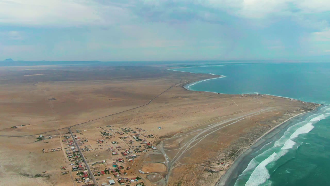 Aerial frame over Punta Abreojos, Baja California Sur, showing a small desert settlement at lower left, white surf on the right shoreline, open flats inland, and no visible people.