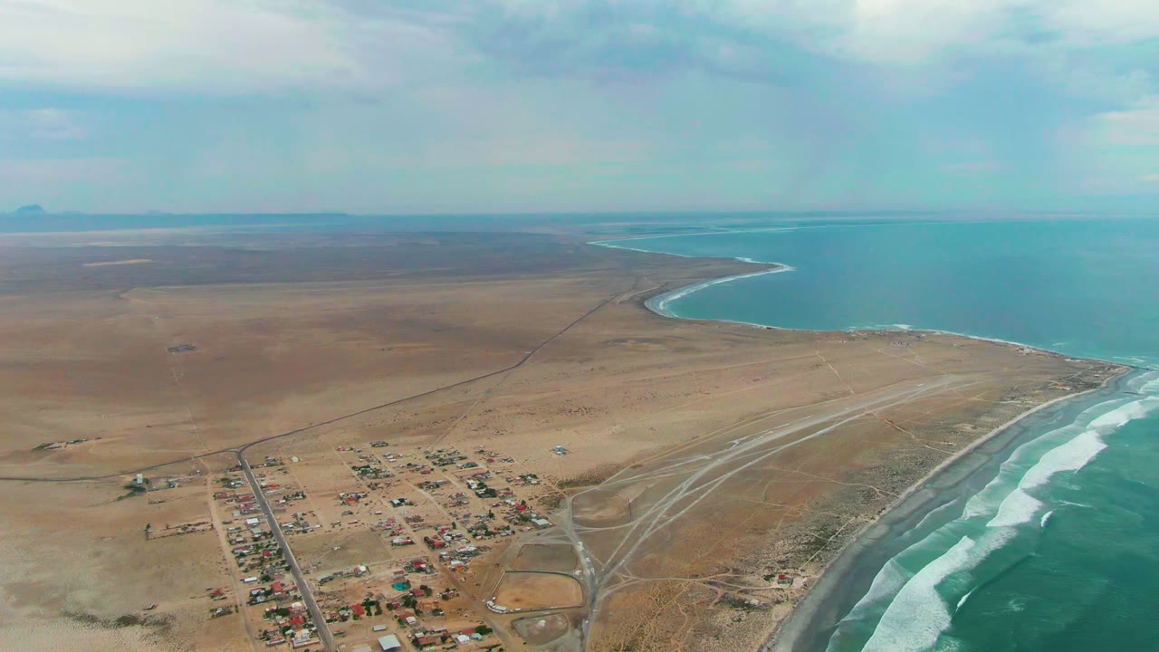 Wide aerial coastline with settlement, sandy roads, airstrip, and ocean waves