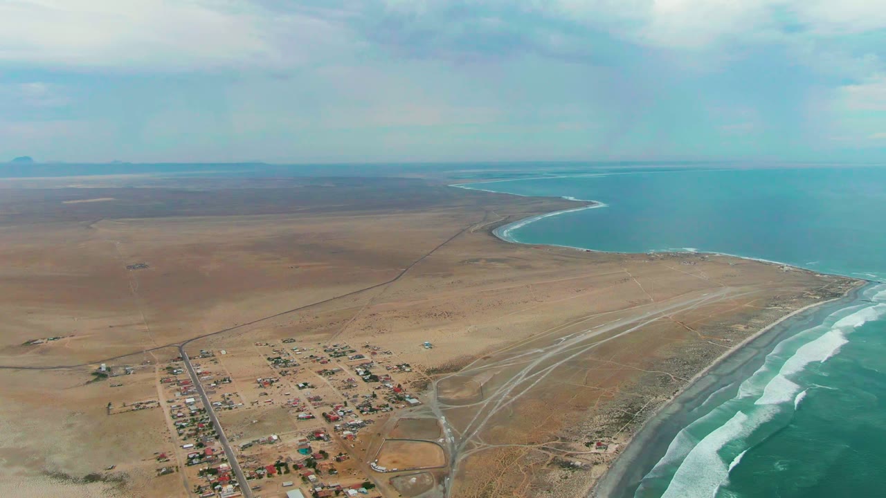 High aerial frame over Punta Abreojos showing brown arid land, a compact cluster of buildings near the bottom, surf bands along the right edge, blue water beyond, and no visible people.