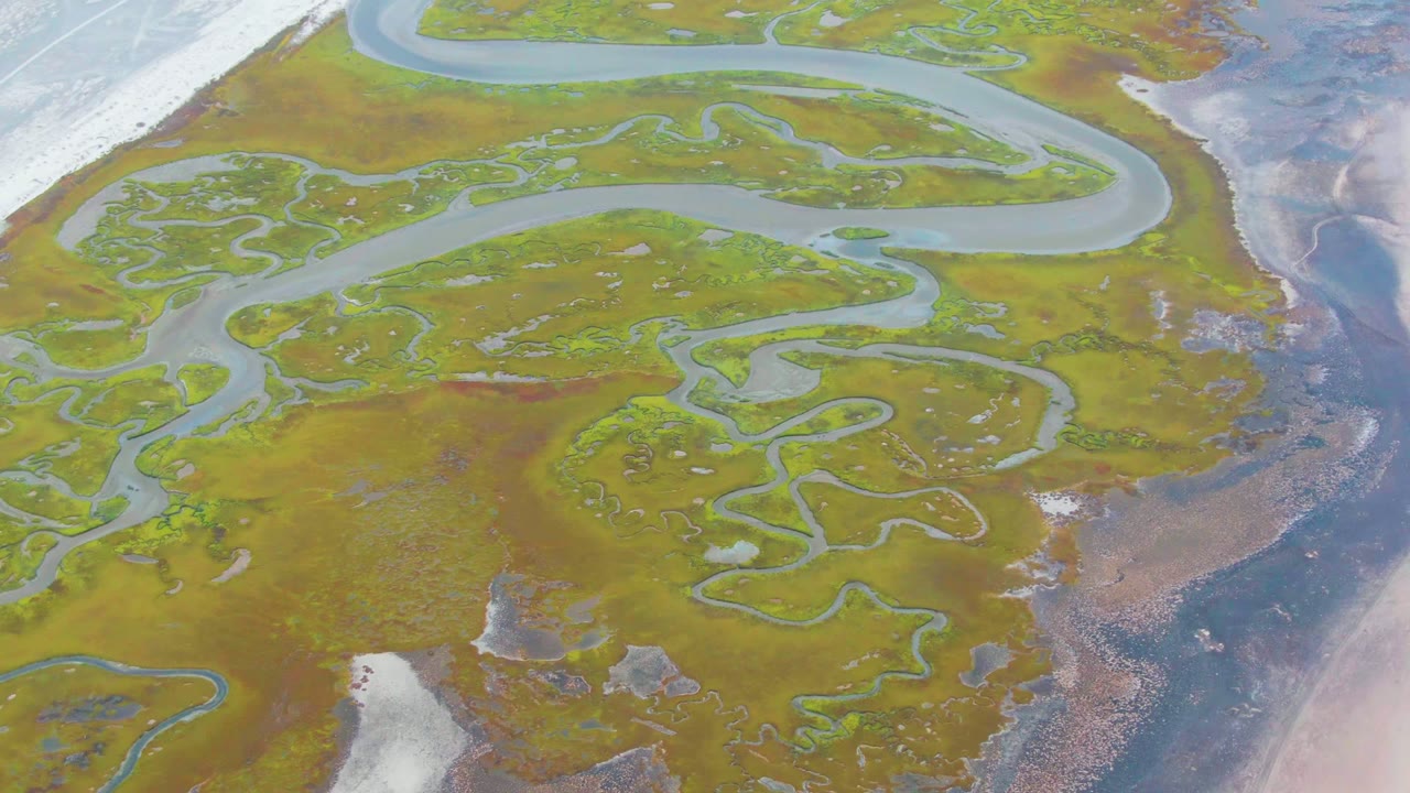 Aerial view over wetland waterways in Punta Abreojos, Baja California Sur, with green vegetation, brown earth, white snow or ice patches, and no visible people.