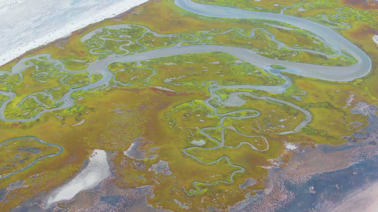 Top-down Punta Abreojos wetland frame showing looping blue-green channels across ochre and green marsh, white ice or snow along the upper left edge and lower left patch, and no visible people.
