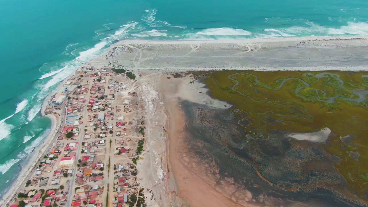 Opening aerial frame over Punta Abreojos, Baja California Sur, showing turquoise surf, a pale sandy shore, red-roofed houses, and green-brown wetland with no visible people.