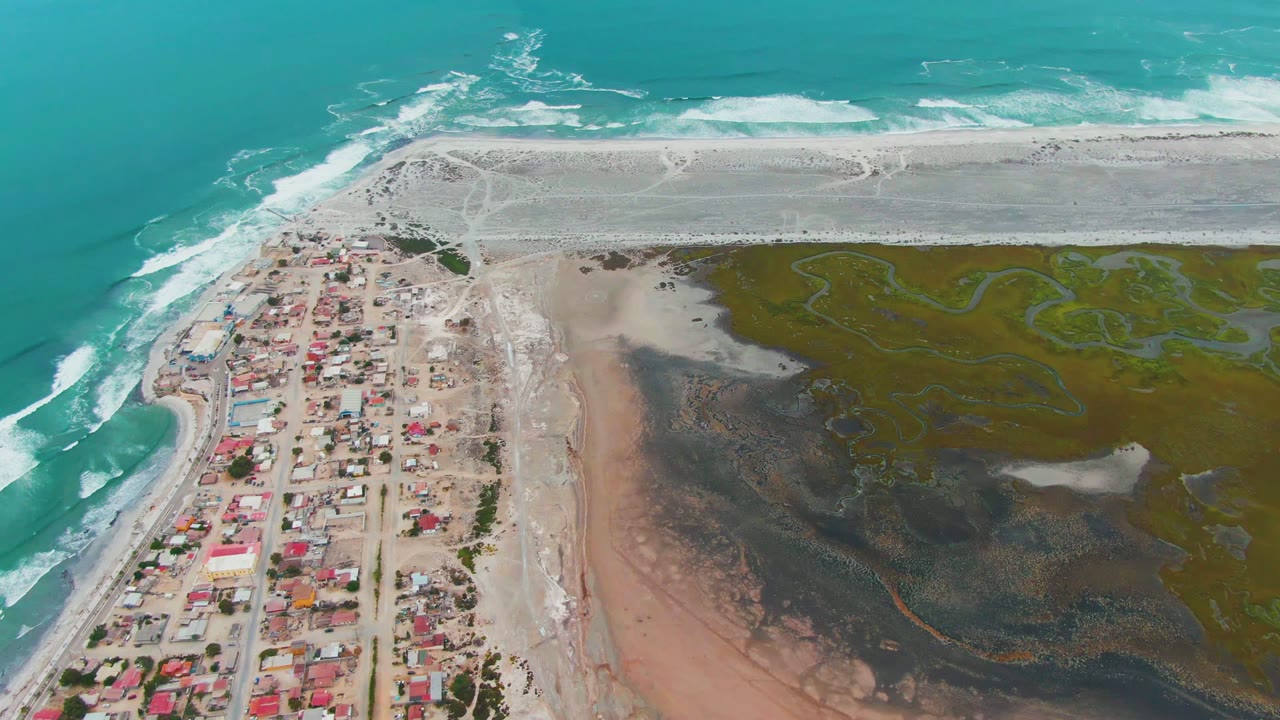 Aerial frame over Punta Abreojos with the town filling more of the lower-left edge, ocean waves breaking along the beach, and marsh channels spread across the right side, with no visible people.