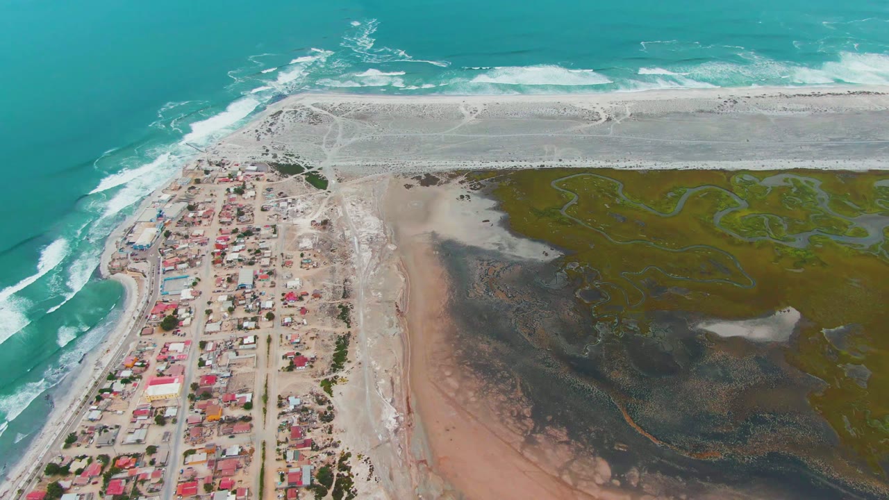 Punta Abreojos aerial view with white wave bands across the upper shore, compact homes below, and a large wetland basin to the right, with no visible people in the frame.