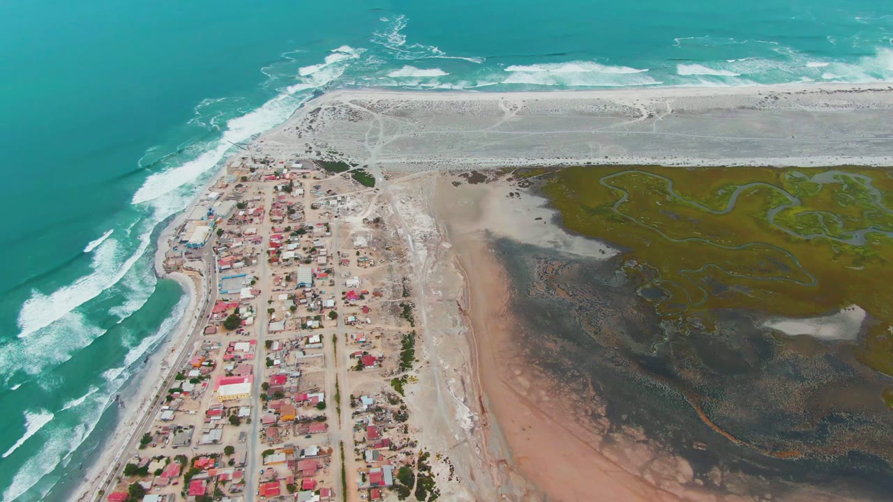 Aerial frame over Punta Abreojos, Baja California Sur, with turquoise ocean surf on the left, red-roofed village blocks near the beach, tidal flats to the right, and no visible people.