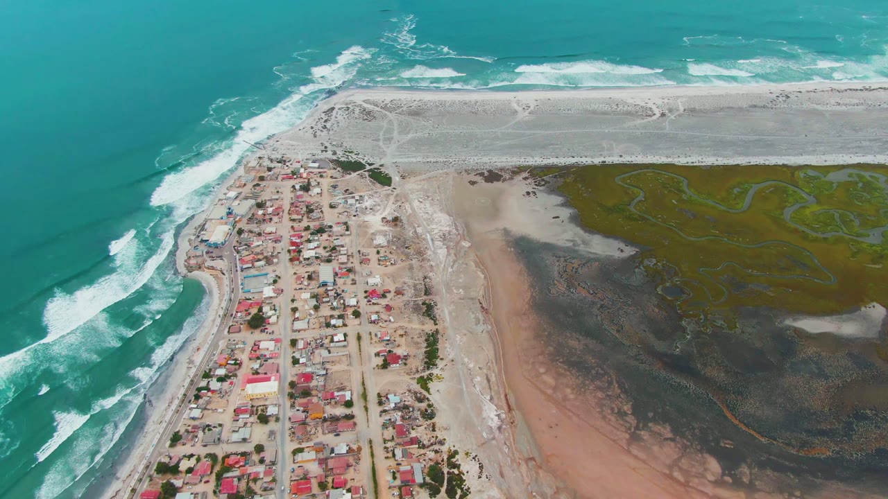 High aerial frame of Punta Abreojos with the village grid below center, teal water and foam on the left, pale sandbar shoreline beyond, green marsh channels on the right, and no visible people.