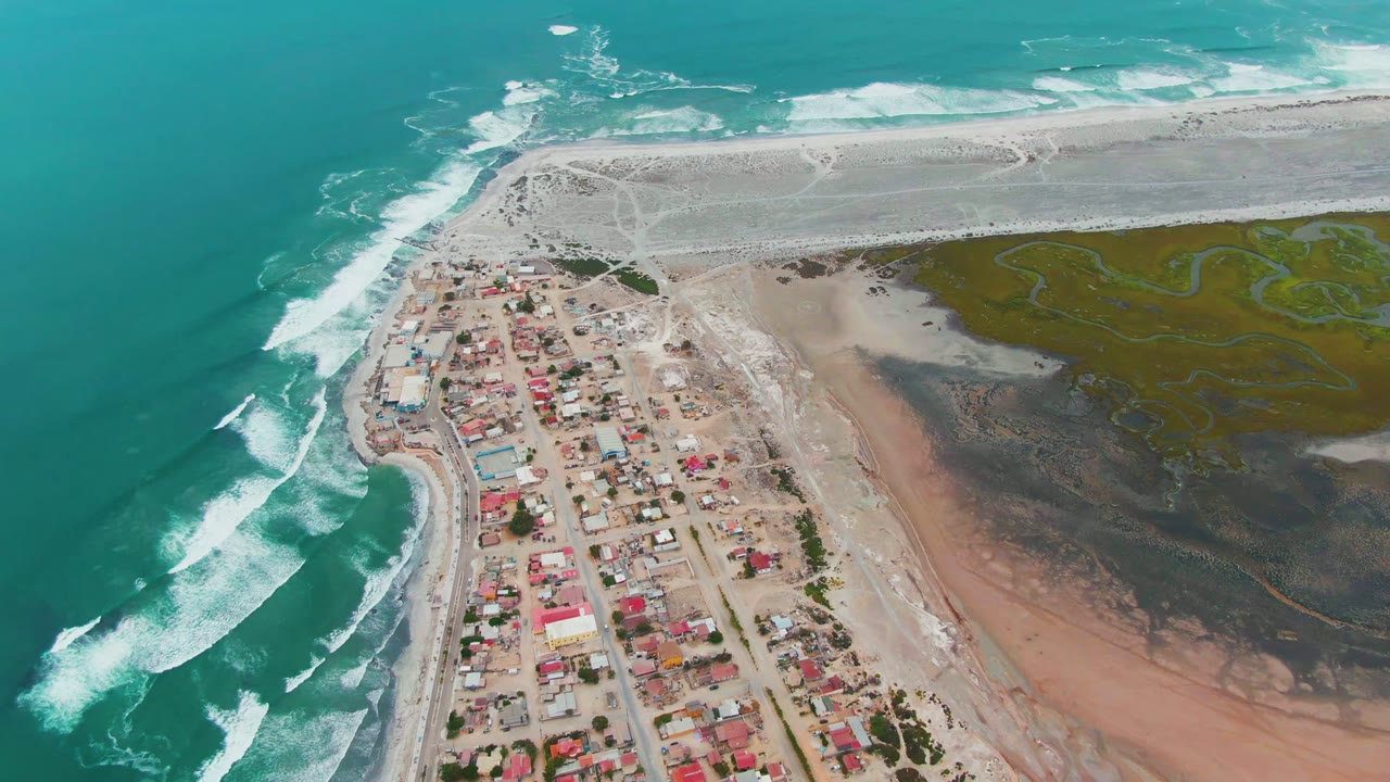 Aerial frame over Punta Abreojos showing turquoise water and white surf on the left, a compact coastal village below center, pale beach above, marshland to the right, and no visible people.