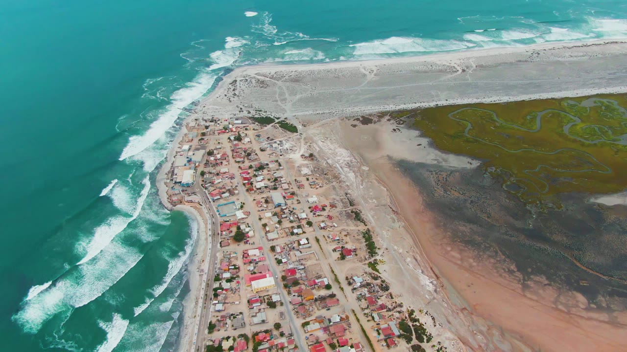 Drone view of surf lines beside a coastal village and marsh flats.