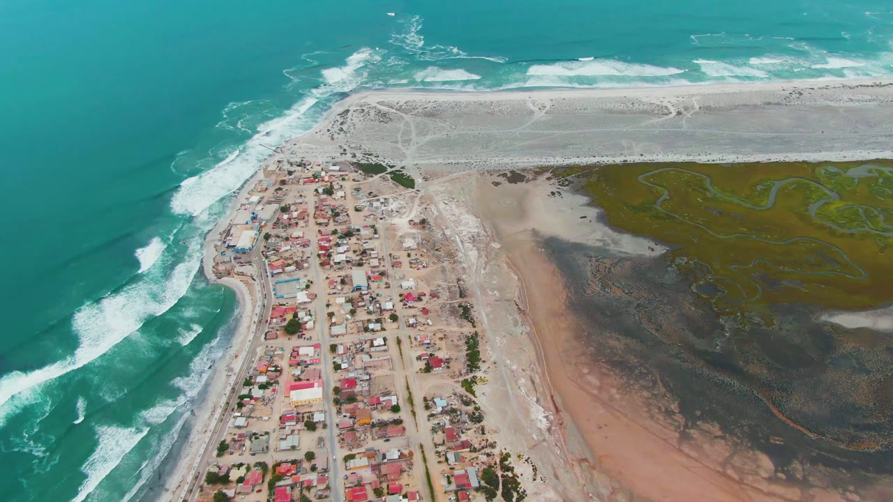 Aerial view over Punta Abreojos BCS Mexico with turquoise surf on the left, a compact village in the center, muddy tidal flats on the right, and no visible people.