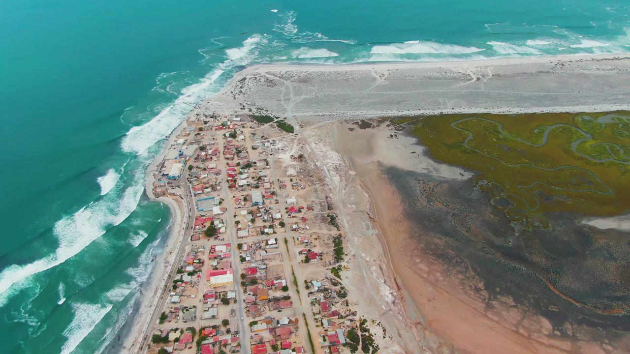 Coastal settlement beside white surf and dark tidal mud from above.