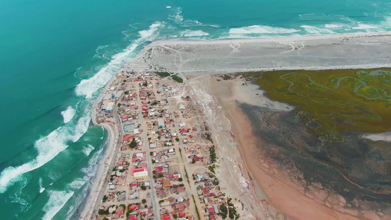 Drone-height view over Punta Abreojos BCS Mexico with ocean breakers curling along the left beach, clustered homes below, muddy lagoon-like shore to the right, and no visible people.