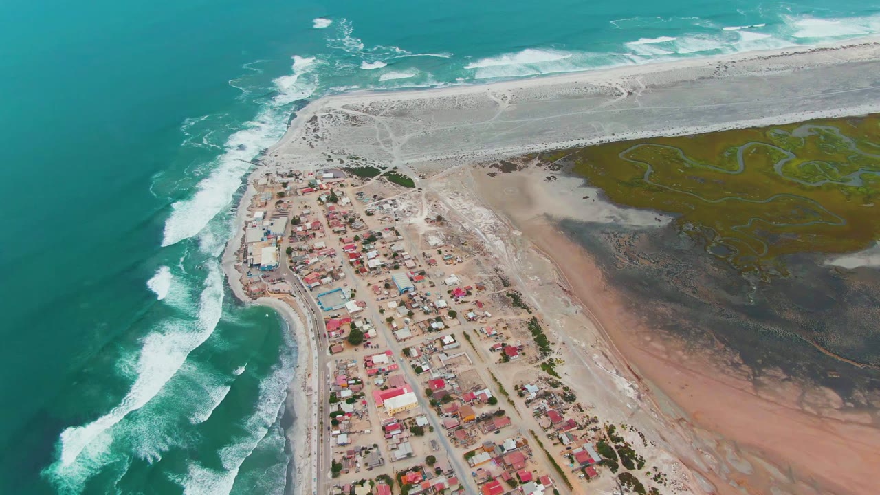 Aerial frame over Punta Abreojos, Baja California Sur, with turquoise surf on the left, red-roofed houses near the beach, a pale sandy jetty above, green lagoon flats to the right, and no visible people.