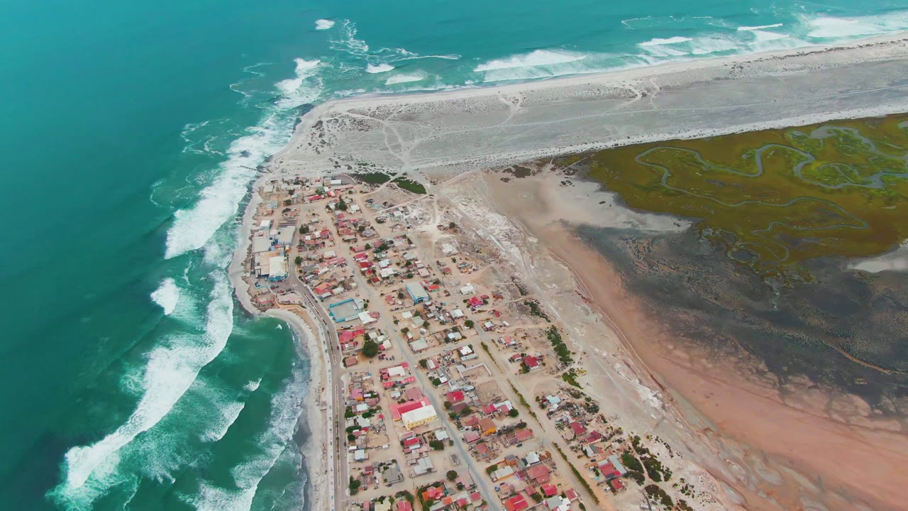 Drone view of surf beside a small beach settlement and sandy spit.