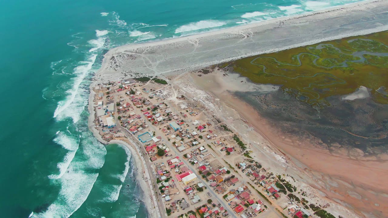 Opening aerial frame over Punta Abreojos, Baja California Sur, with turquoise surf on the left, clustered homes below, tidal flats to the right, and no visible people.