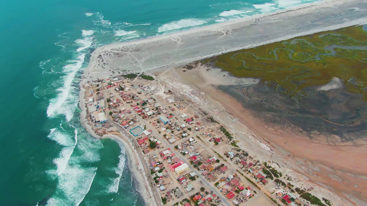 Mid-clip aerial over Punta Abreojos showing bright surf curling at the left edge, compact houses near the lower middle, a pale sand bar above, and no visible people.