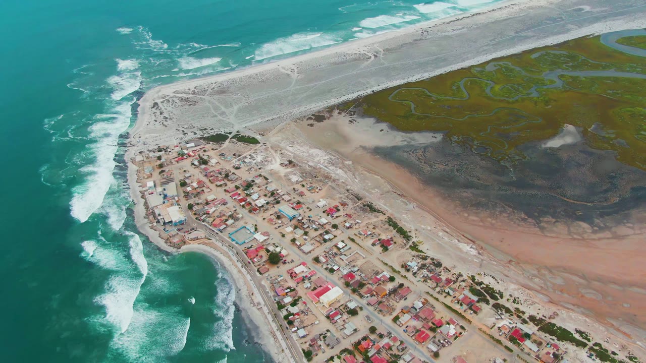 High aerial frame over Punta Abreojos, Mexico, with turquoise surf breaking along the left edge, clustered village buildings below, pale sand flats above, green tidal channels to the right, and no visible people.