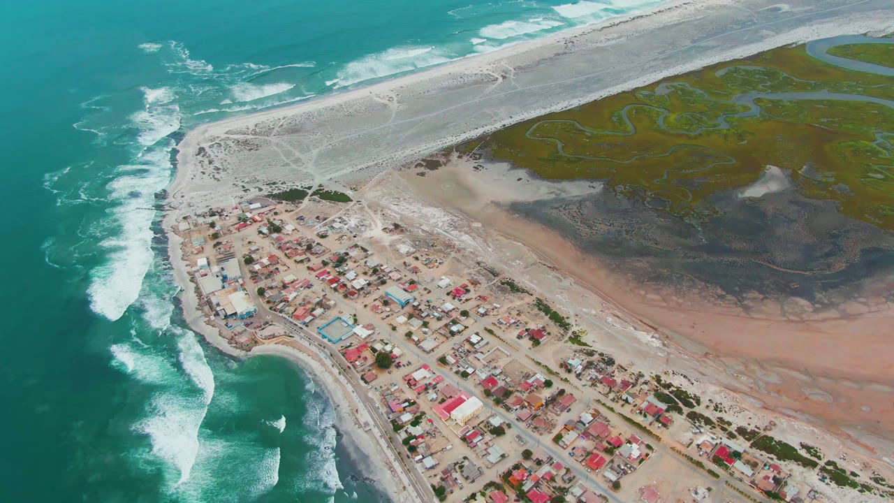 The drone angle holds over Punta Abreojos as foamy waves advance along the left shoreline, red and light-colored roofs cluster near the road, and pale sandbars meet green channels inland, with no visible people.