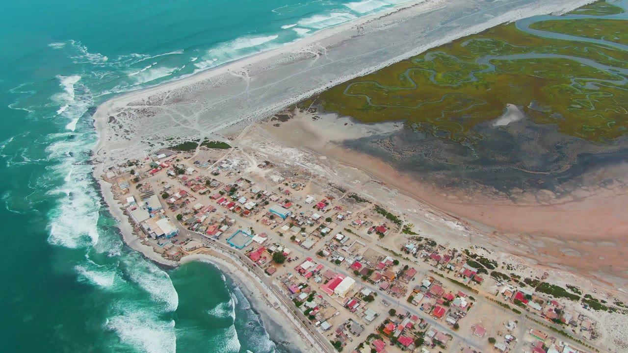 Aerial frame over Punta Abreojos, Baja California Sur, with turquoise surf on the left, red-roofed buildings below, pale beach across the center, green lagoon channels above, and no visible people.