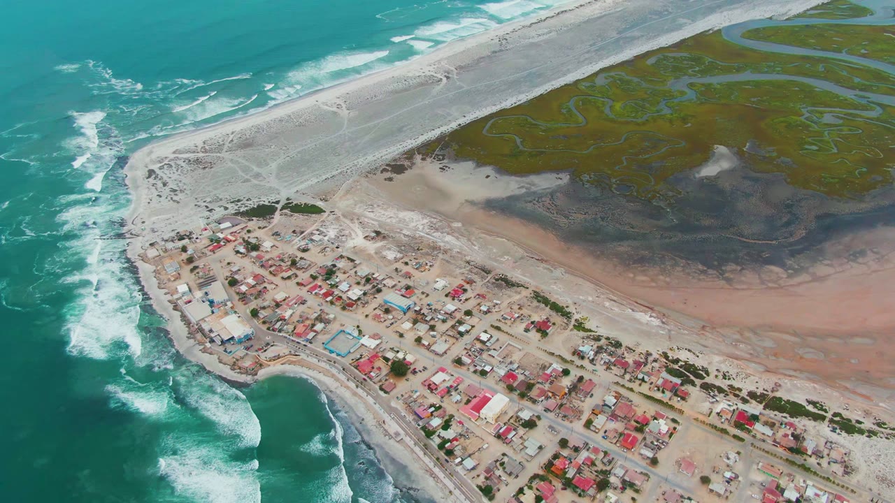 Drone view of waves breaking beside a small coastal settlement and lagoon flats.