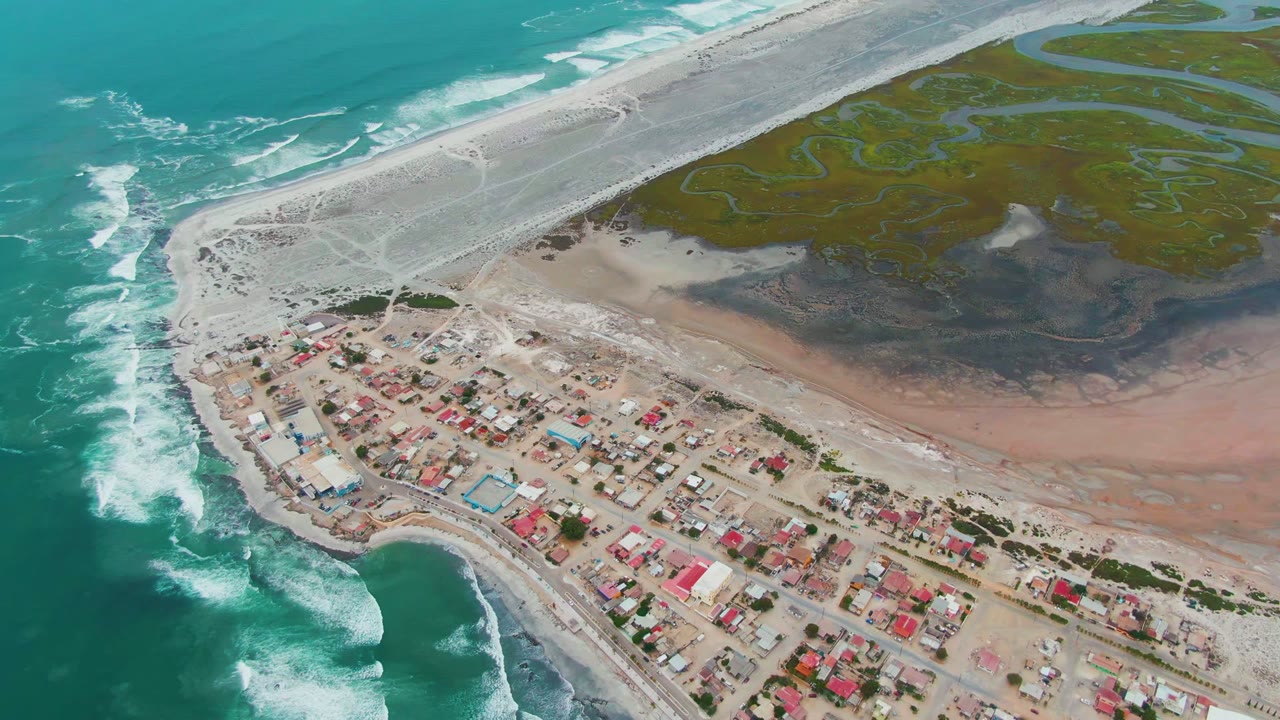 High aerial over Punta Abreojos showing foam lines advancing along the ocean side, red-roofed buildings near the lower shore, pale sand bars through the center, and green lagoon water in the background with no visible people.