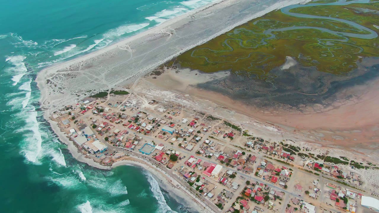 Aerial view over Punta Abreojos, Baja California Sur, showing turquoise surf at left, a compact coastal village below, sandy tracks, green wetland channels, and no visible people.