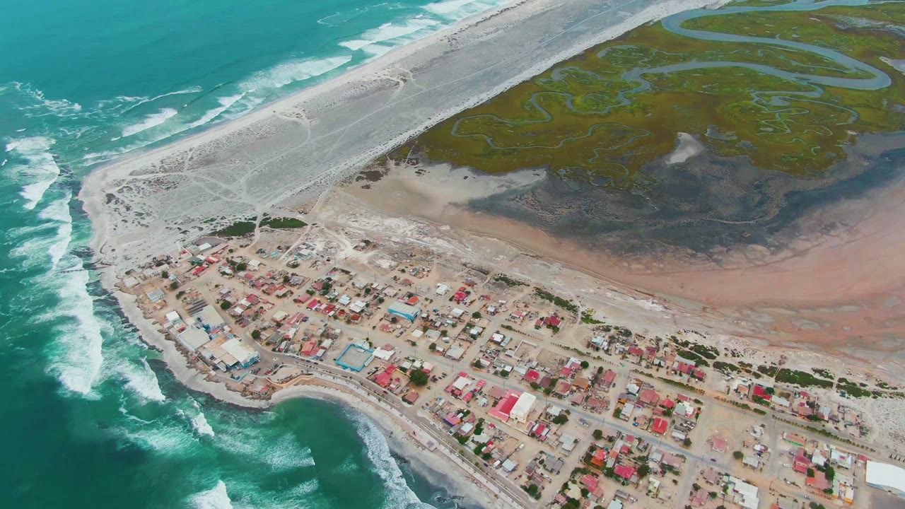 Drone view of surf, beach road, village rooftops, and marsh channels.