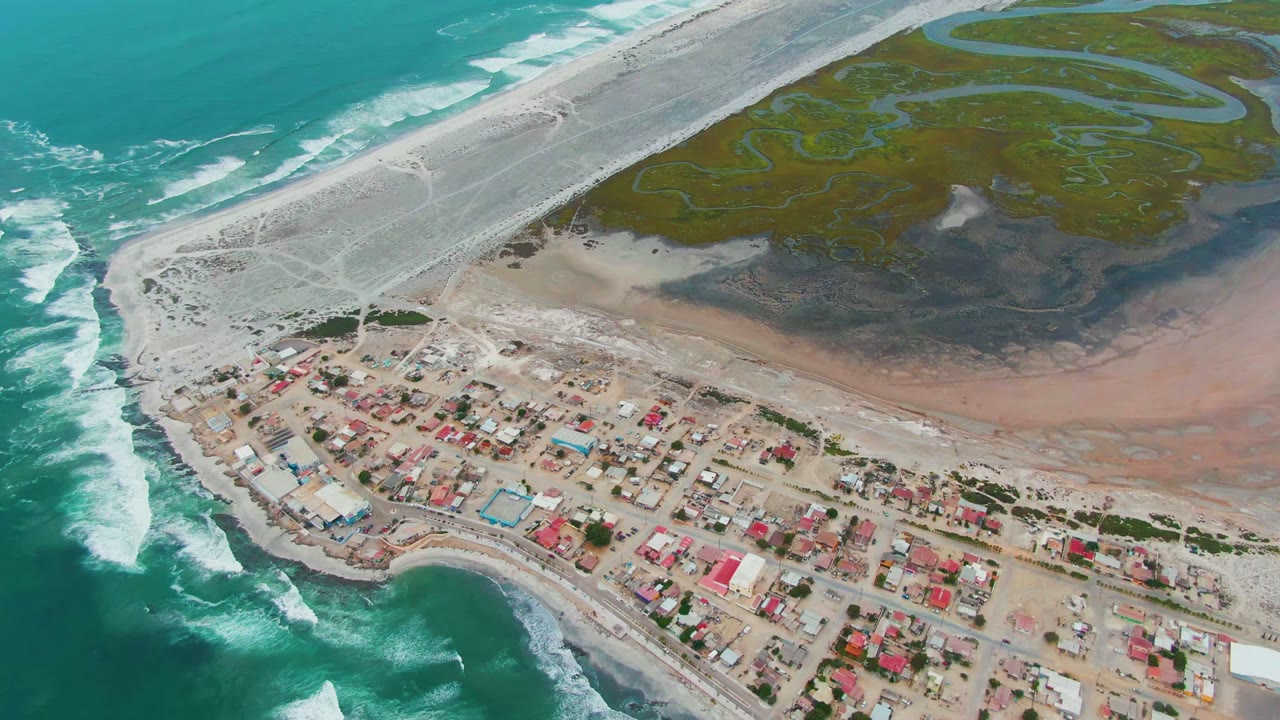 High aerial frame of Punta Abreojos showing stronger white surf patterns at left, the shoreline settlement across the bottom, pale sand flats in the center, green wetlands at top, and no visible people.