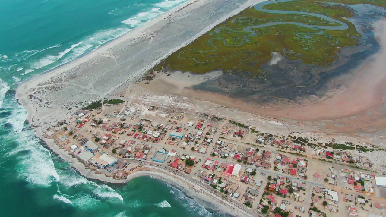 Aerial view over Punta Abreojos with turquoise surf at left, red-roofed village buildings below, a pale causeway crossing the beach area, green marsh channels inland, and no visible people.