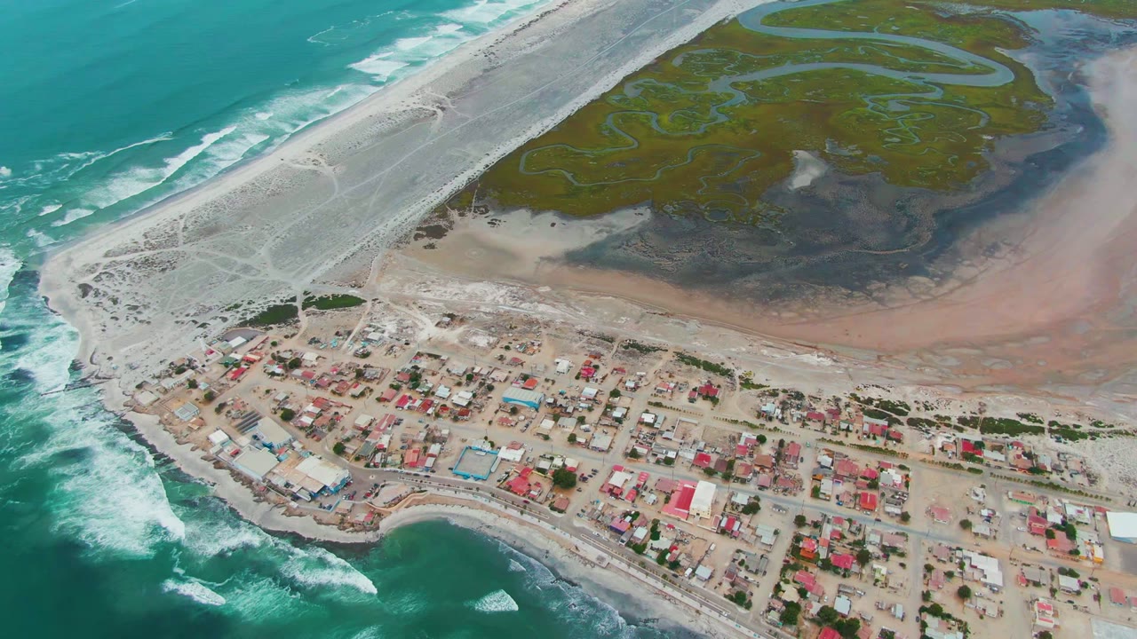 Coastal village beside white surf, sandy shore, diagonal causeway, and green marsh.