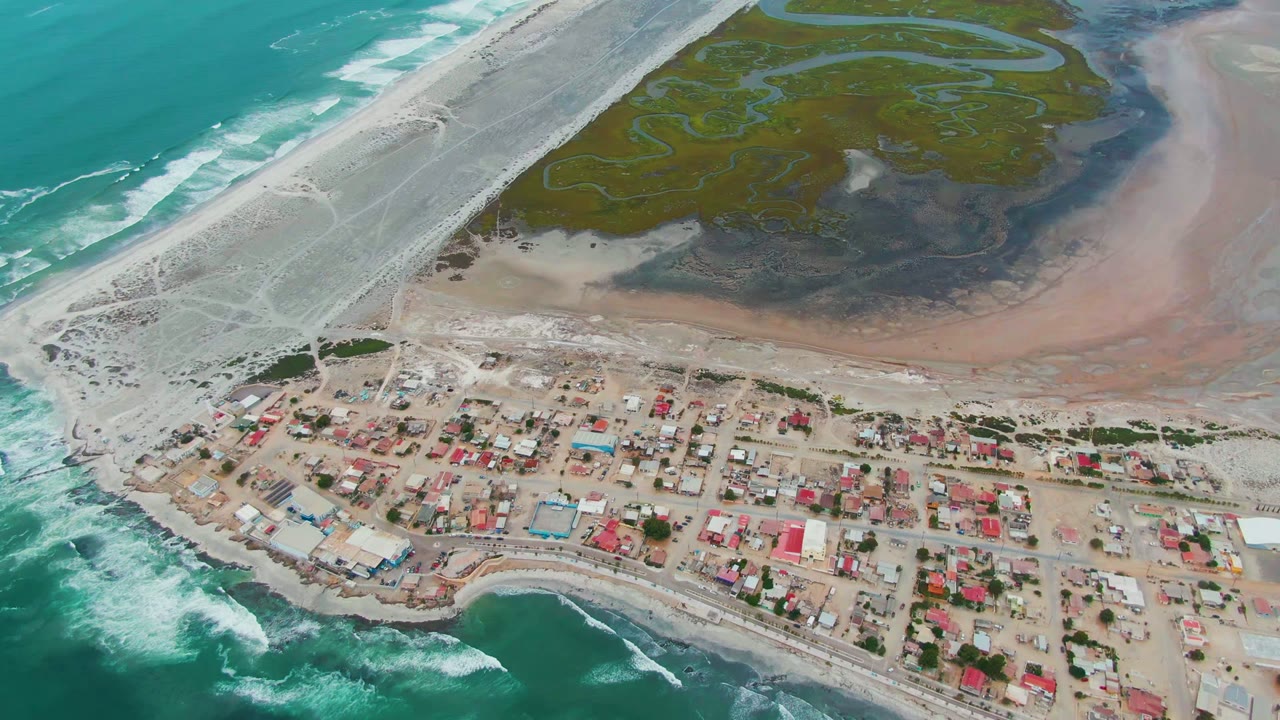 Aerial view over Punta Abreojos, Baja California Sur, with turquoise surf at the lower left, village roofs across the shore, lagoon marsh above, and no visible people.