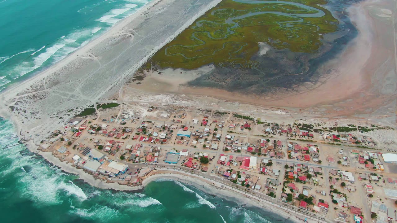 Drone view of shoreline village with red roofs, surf, and lagoon.