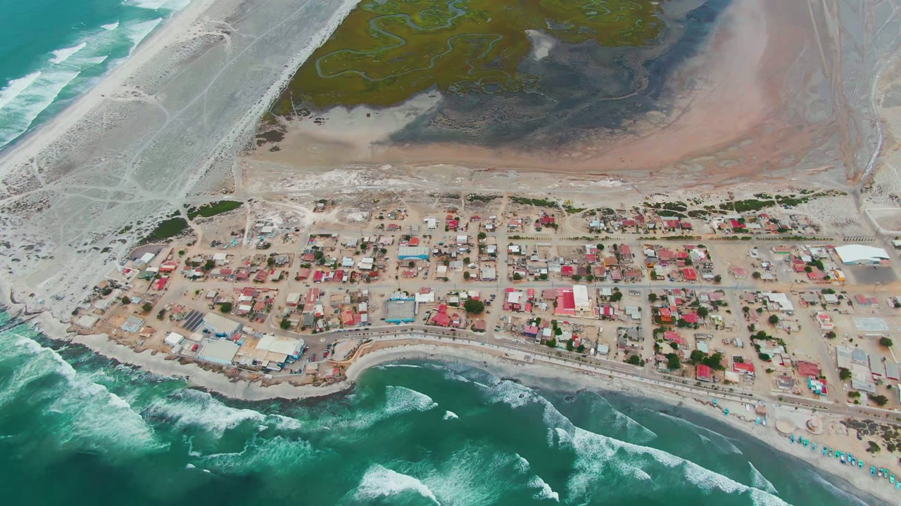 Opening aerial view of Punta Abreojos, Baja California Sur, with turquoise surf at the lower edge, a sandy village in the center, lagoon flats behind it, and no visible people.