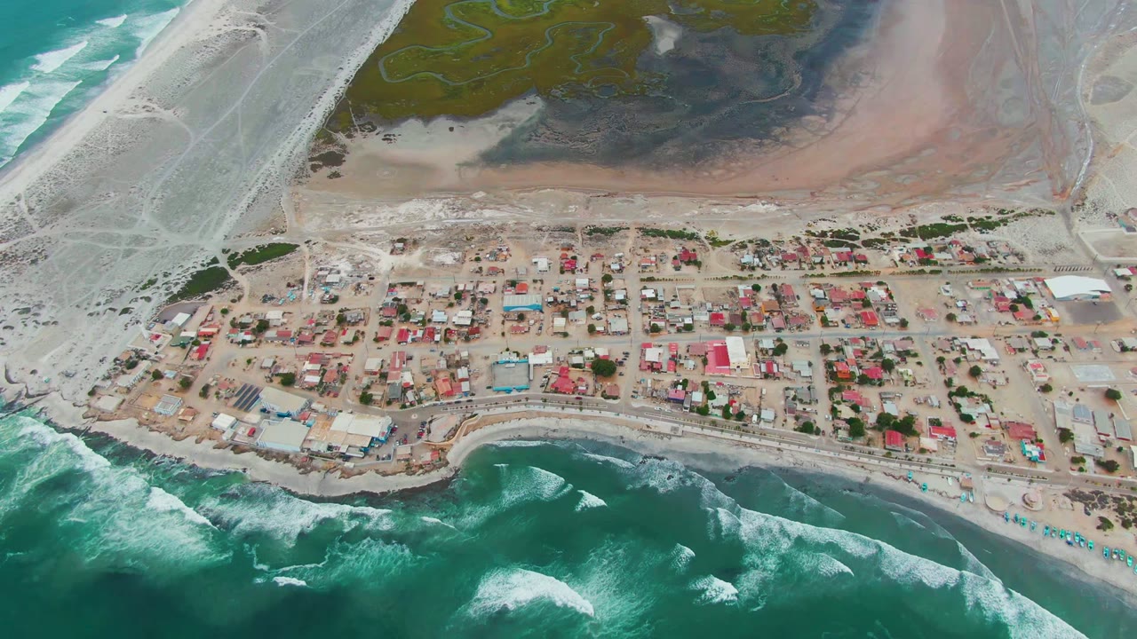 Mid-clip aerial view over Punta Abreojos with rows of small coastal buildings, white wave breaks along the lower beach, lagoon channels in the upper background, and no visible people.