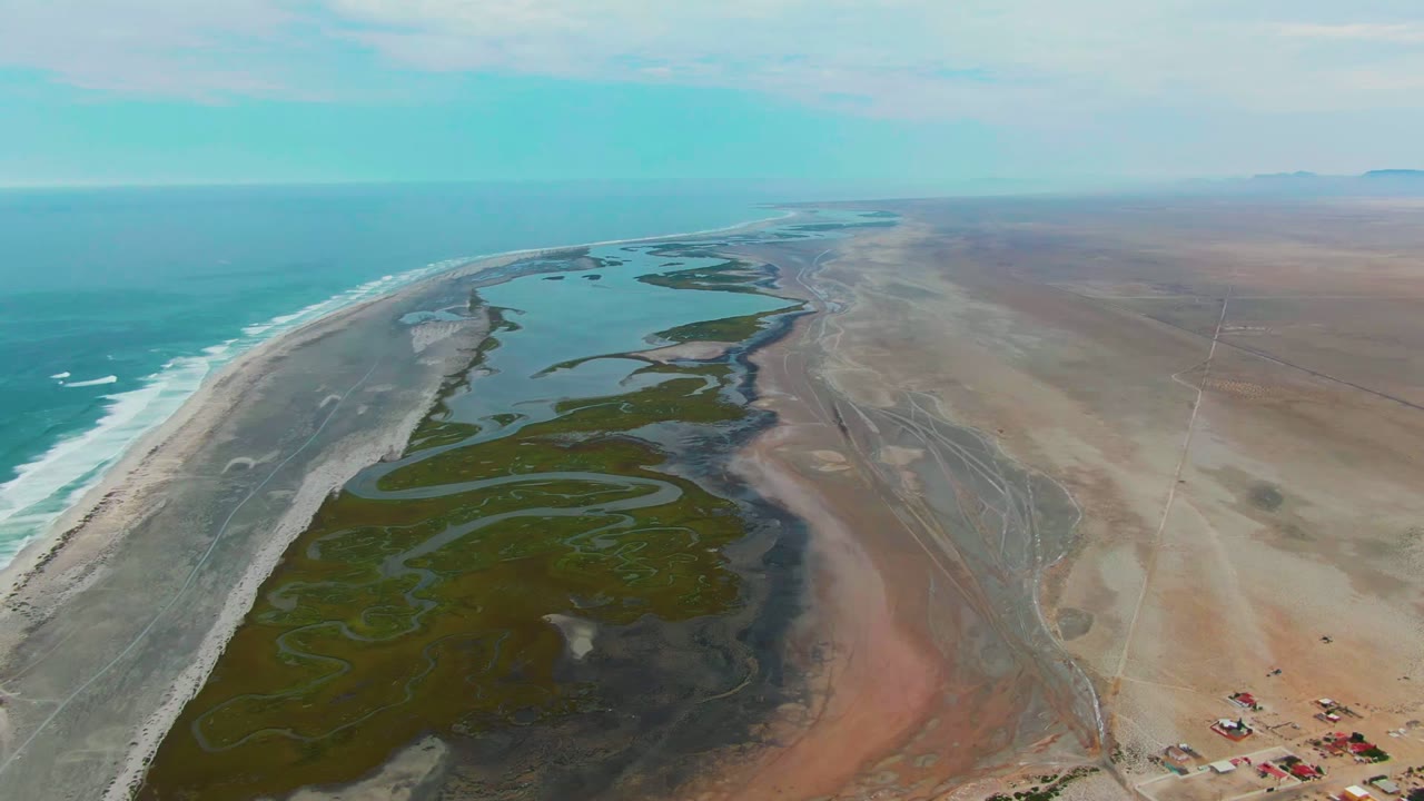 Aerial view at Punta Abreojos, Baja California Sur, showing turquoise ocean, white surf, pale beach, green lagoon channels, tan desert flats, and no visible people.