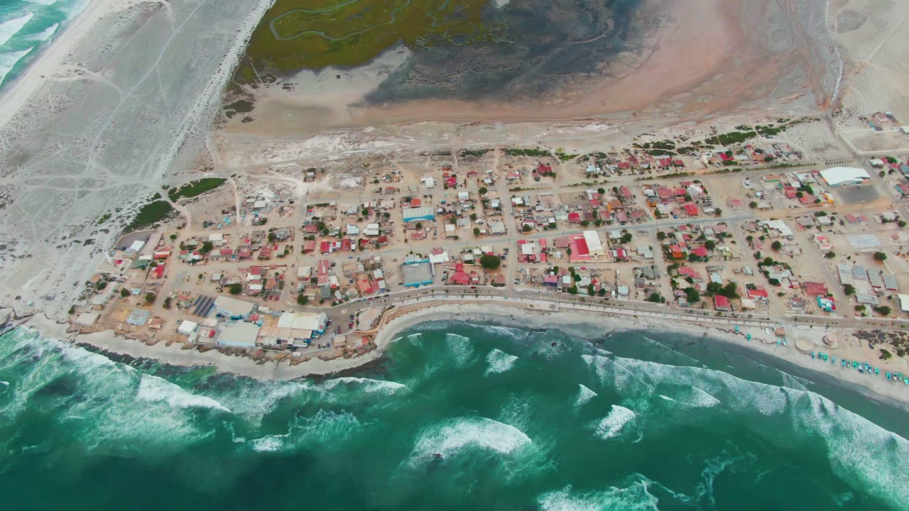 Overhead view of Punta Abreojos in Baja California Sur, Mexico, with turquoise surf below the beach, red and blue roofs across the settlement, pale sand flats behind town, and no visible people.