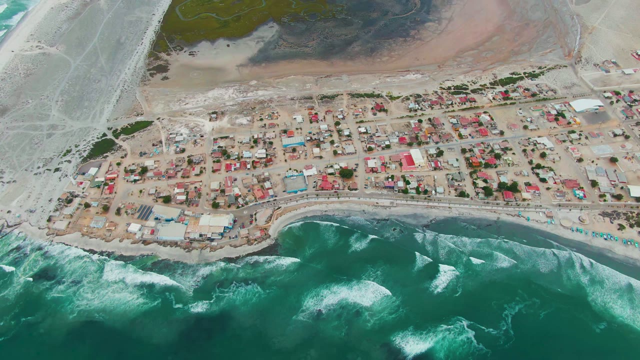 High overhead view of Punta Abreojos with whitecaps spreading through the turquoise water, a narrow beach strip, clustered red and blue-roofed buildings, light-colored roads, and no visible people.