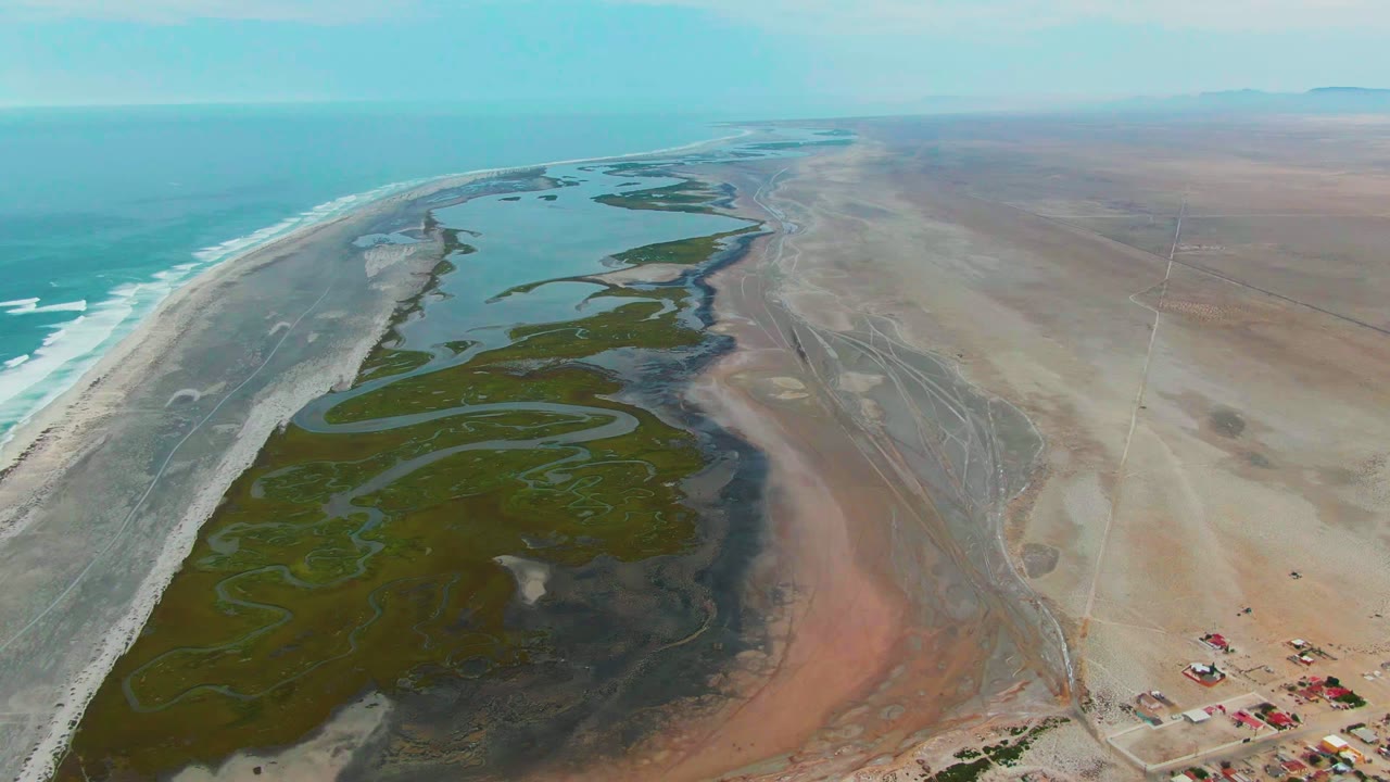 Wide aerial frame over Punta Abreojos, Baja California Sur, showing turquoise ocean, pale beach, green wetland channels, dry brown flats, small structures at the lower right, and no visible people.