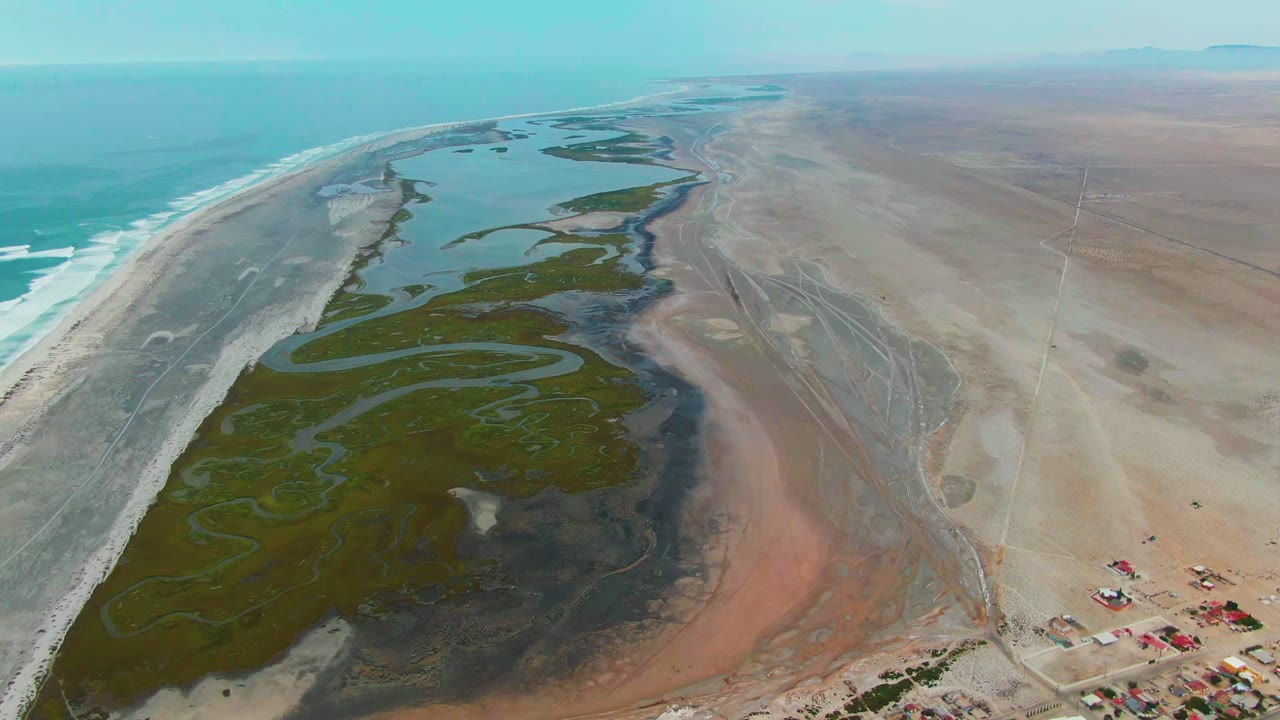 High aerial frame at Punta Abreojos showing the blue-green ocean, long sandy shoreline, dark green wetland patches, exposed flats, a few small lower-right structures, and no visible people.